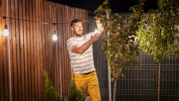 Party time in backyard with happy millennial man hanging string lights in trees – Weekend summer night mood with smiling guy arranging the light garland for outdoor dinner party in home garden
